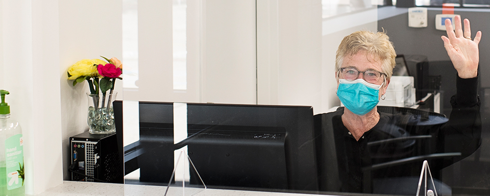 Woman waving while wearing glasses and a surgical mask seated at a computer behind a protective shield.