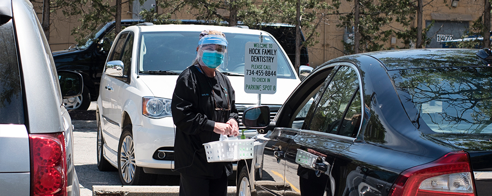 Woman in scrubs, surgical mask, and face shield standing outside of a black car.